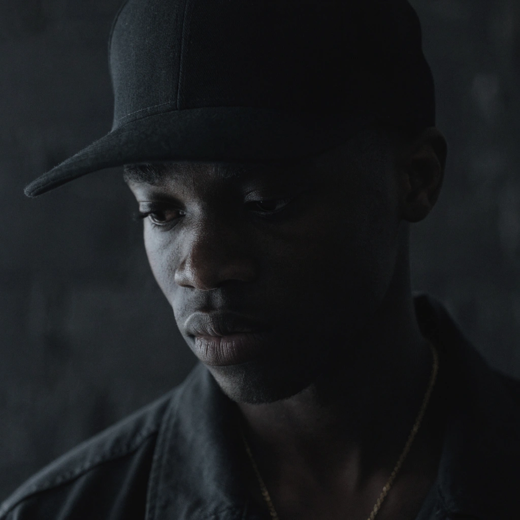 Person wearing a plain black baseball cap in a dark, minimalist portrait
